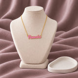 Script Name Enamel Necklace — on an ivory display with mauve fabric and white styling props