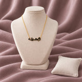Arabic Mum Nickname Enamel Necklace — on an ivory display with mauve fabric and white styling props