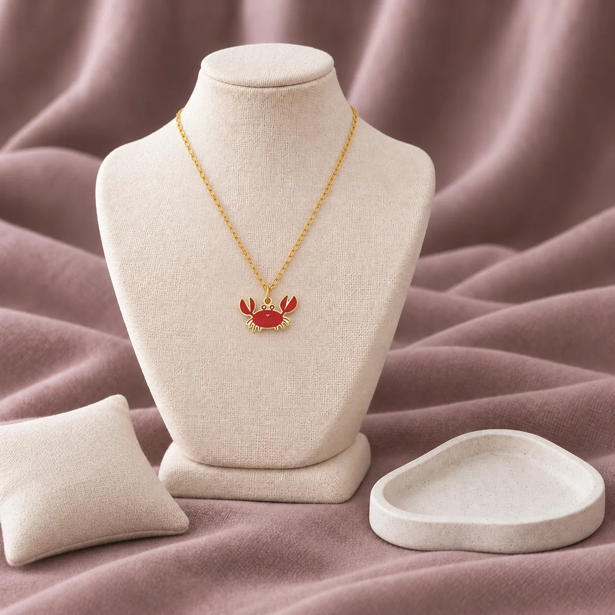 Cutie Crab Enamel Necklace — on an ivory display with mauve fabric and white styling props