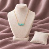 Arabic Block Name Enamel Necklace — on an ivory display with mauve fabric and white styling props